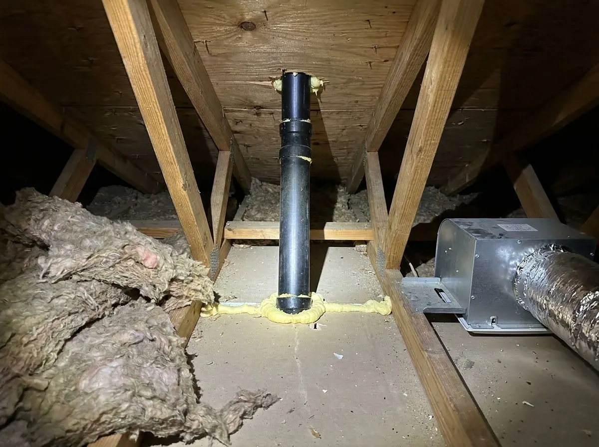 In a cleared attic, a plastic plumbing vent pipe passing through the ceiling has a neat collar of spray foam sealing the gap around it.