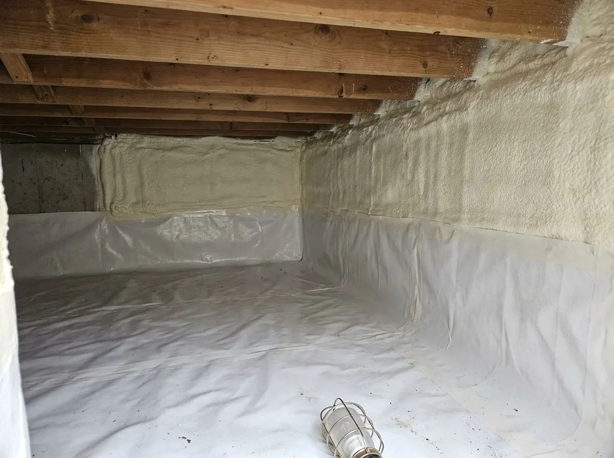 A low crawl space with a heavy white liner covering the ground and lower walls, pale spray foam on most of the upper walls, and one short stretch of bare concrete still to be foamed.