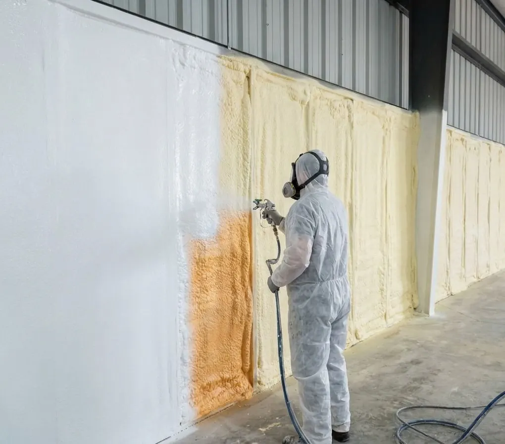 An installer in a respirator sprays a white coating over yellow spray foam on the walls of a steel-clad shop, turning the foam white section by section.
